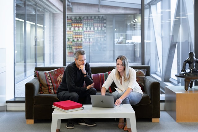 A man and a woman sitting on a couch in an office and looking at a laptop.