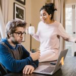 Upset woman holding a coffee mug sitting on a desk and trying to argue with a man who's looking at his laptop